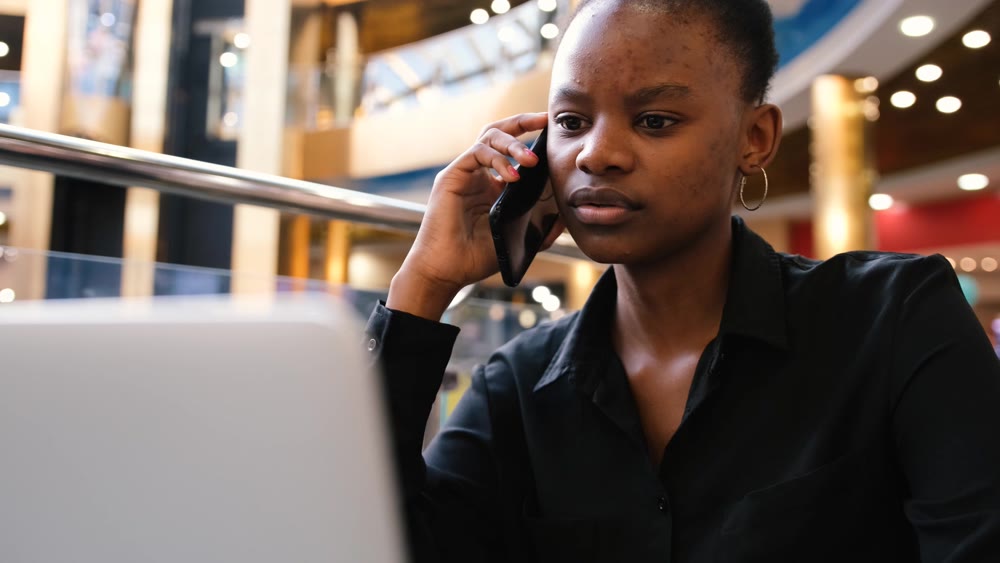An African woman works at a computer and talks on the phone while typing on the keyboard.
