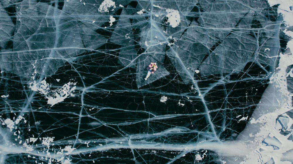 A tourist girl in bright clothes and sunglasses smiles happily, lies on the frozen blue ice of Lake Baikal and controls the drone up. An incredible pattern of crack bubbles. Siberia Russia tourist attraction.