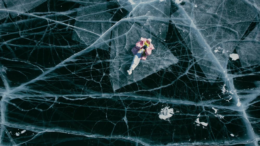 A tourist girl in bright clothes and sunglasses smiles happily, lies on the frozen blue ice of Lake Baikal and controls the drone up. An incredible pattern of crack bubbles. Siberia Russia tourist attraction.